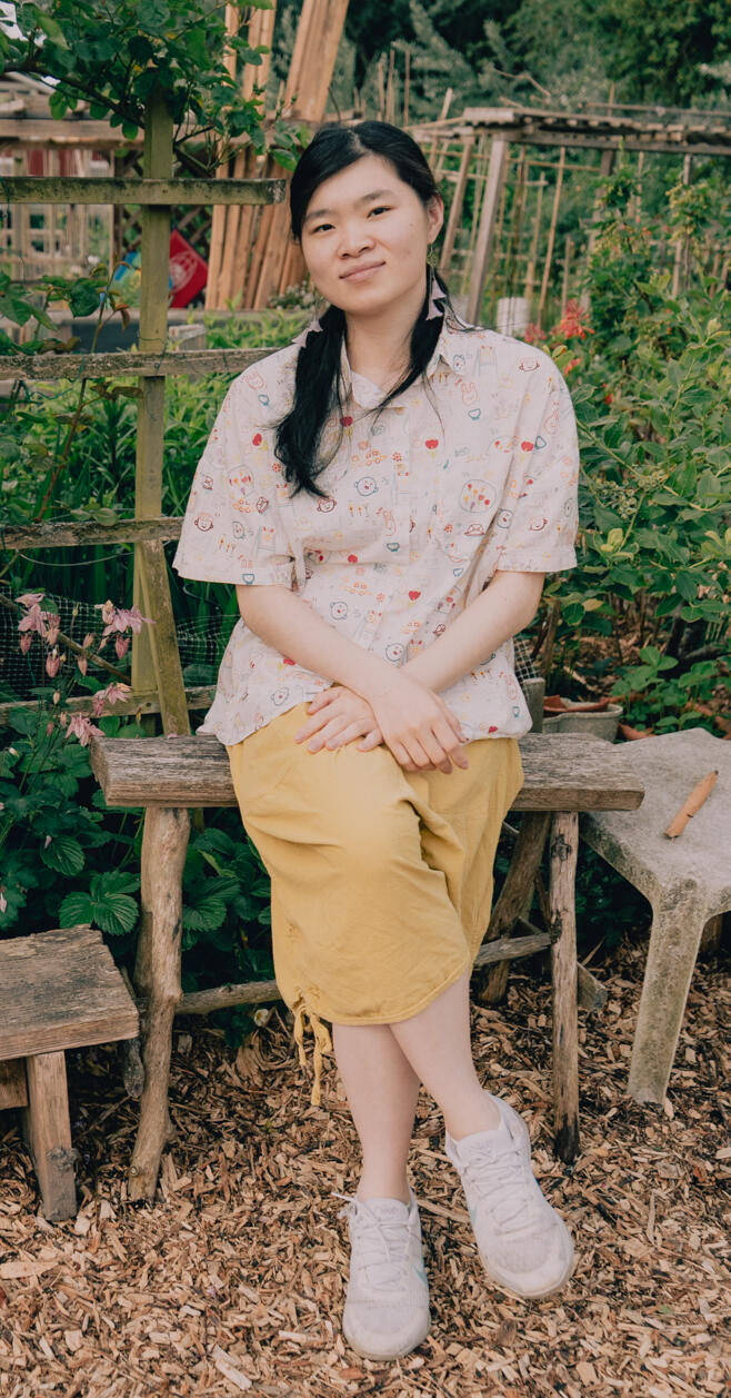 photographer: Ashley Cheng (IG: @0922s9) Photo of the artist, an asian woman wearing a dangling pink flower earrings, a white short-sleeve button up with a colourful doodle pattern on it, yellow-green shorts, and white sneakers. She is seated on a small driftwood bench with a sprawling garden beh